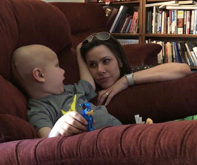 Jacob sits on a red couch holding a small blue toy and looks toward his mom, who is resting beside him.