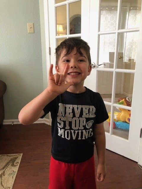 A smiling child in a black Never Stop Moving shirt holds up an I love you hand sign in a living room.