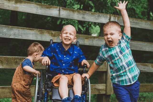 Jacob laughs from a wheelchair outdoors while one sibling leans in and another raises an arm, in front of a wooden fence.