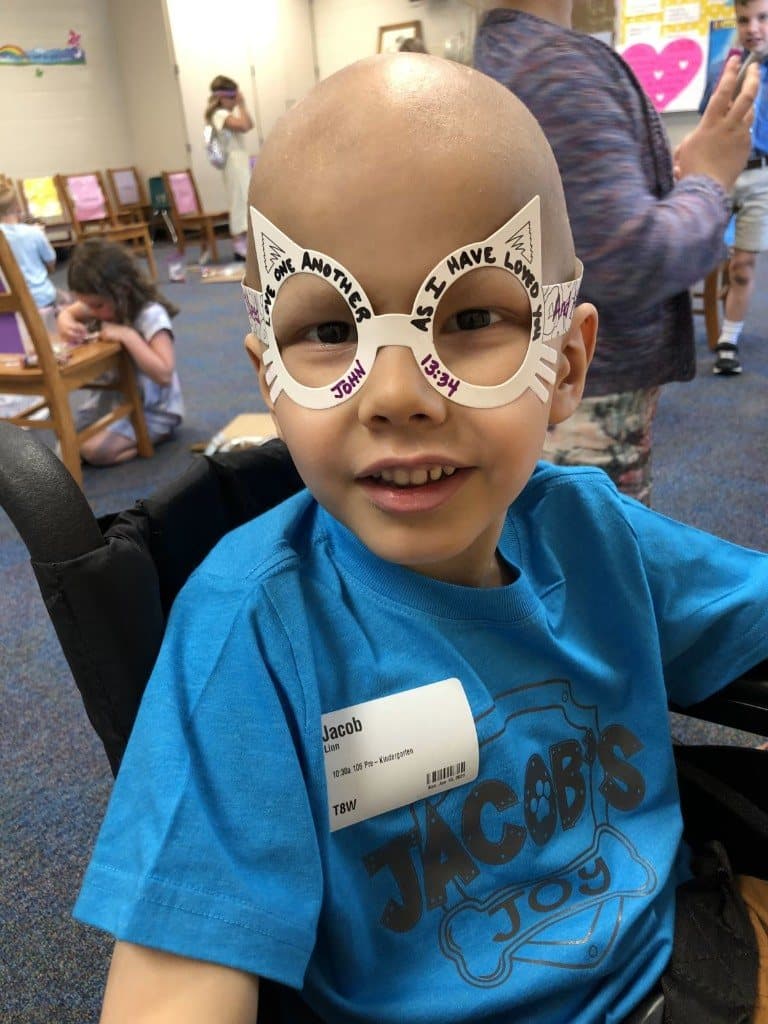 Jacob smiles wearing oversized paper glasses painted with the words love one another, John 13:34, and a bright blue Jacob’s Joy shirt.