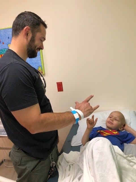 Jacob in a Superman shirt lies in a hospital bed and returns an I love you hand sign to his dad, who stands beside him smiling.