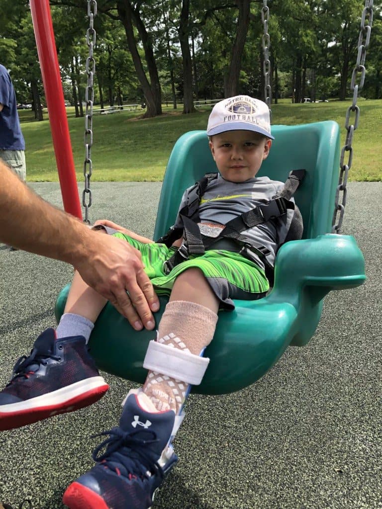 Jacob sits buckled into a green park swing, wearing a white Notre Dame football cap and bright green shorts.